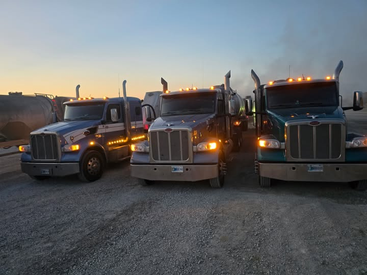 Three Ulyat Trucking Peterbilt semi tractors lined up on a gravel lot with their headlights on